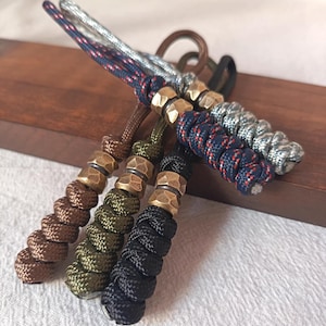 Op de afbeelding: Een verzameling handgemaakte sleutelhangers met gevlochten koord en messing accenten. De sleutelhangers hebben verschillende kleuren, waaronder bruin, olijfgroen, zwart, marineblauw en zilver. Elke sleutelhanger heeft een uniek ontwerp en textuur.