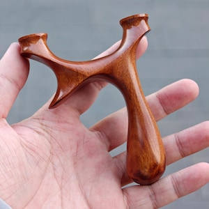 May include: A handcrafted wooden slingshot with a smooth, polished finish. The slingshot is made of a rich, reddish-brown wood and has a classic Y-shape design. The handle is rounded for a comfortable grip.