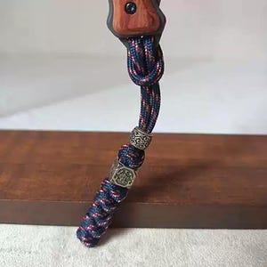 Op de afbeelding: Een koord gemaakt van donkerblauw en rood gespikkeld paracord met een knoop aan de bovenkant, twee zilverkleurige metalen kralen met ingewikkelde reliëfpatronen en een gevlochten ontwerp, bevestigd aan een houten handvat met een enkele zwarte schroef.