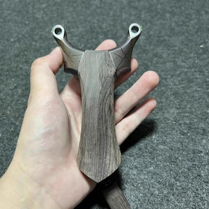 Puede incluir: Una honda de madera con un mango marrón oscuro y un marco metálico. La honda se sostiene en una mano, mostrando su diseño y construcción. El marco tiene un patrón agrietado.