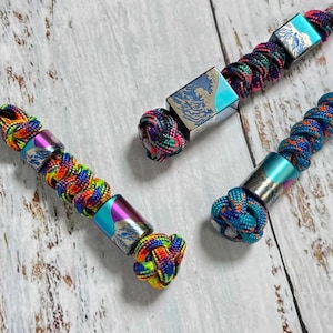 May include: Close-up of three colorful lanyards with decorative metal beads. Each lanyard features a vibrant, braided cord in shades of blue, purple, green, and orange. The metal beads have a wave design. The lanyards are knotted at the ends.