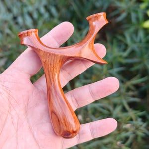 May include: A handcrafted wooden slingshot with a polished finish. The slingshot is made of reddish-brown wood, featuring a curved handle and forked top. The design includes a finger groove for grip and a smooth, glossy surface.