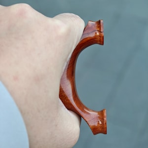 May include: A handcrafted slingshot made of polished reddish-brown wood. The slingshot has a curved handle and a forked top, designed for holding the elastic bands. The wood grain is visible, adding to its natural aesthetic.