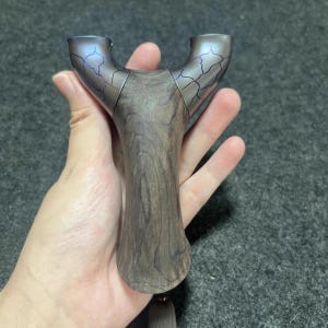 Puede incluir: Una honda con un mango de madera oscura y una parte superior metálica en forma de Y. La parte superior tiene un diseño agrietado azul-púrpura. La honda se sostiene en una mano sobre un fondo gris. El mango es de color marrón oscuro.