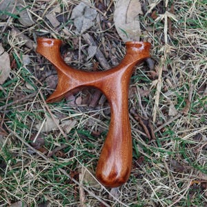 May include: A handcrafted wooden slingshot with a smooth, polished finish. The slingshot is made of reddish-brown wood and has a Y-shaped frame with a rounded handle. It is resting on a bed of dry grass and leaves.