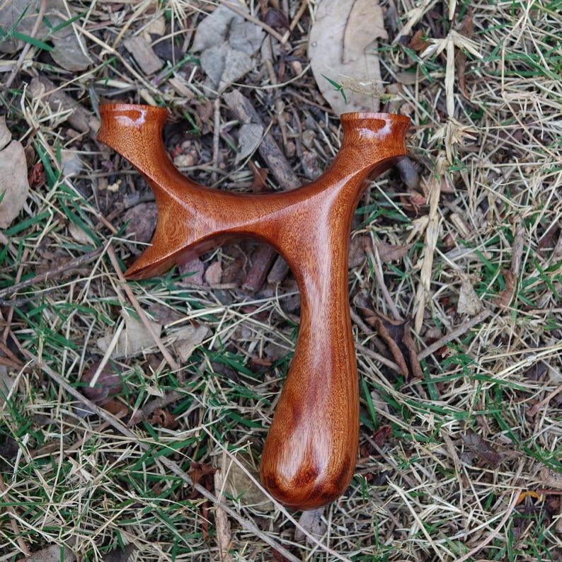 May include: A handcrafted wooden slingshot with a smooth, polished finish. The slingshot is made of reddish-brown wood and has a Y-shaped frame with a rounded handle. It is resting on a bed of dry grass and leaves.