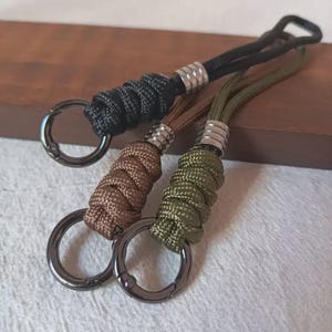 Op de afbeelding: Drie sleutelhangers van gevlochten paracord in zwart, bruin en olijfgroen. Elke sleutelhanger is voorzien van een zilverkleurige metalen kraal, een lus van koord en een ronde, donkergrijze metalen ring met veer voor het bevestigen van sleutels.