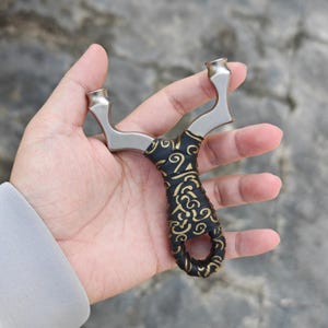 Könnte beinhalten: Eine Steinschleuder mit einem silbernen Metallrahmen und einem schwarzen Griff, der mit einem goldenen, verzierten Muster verziert ist. Die Steinschleuder wird in einer Hand gehalten, um ihr Design hervorzuheben. Die Gesamtästhetik ist eine Mischung aus Funktionalität und dekorativen Details.