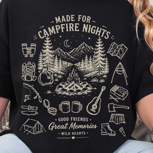 May include: A black short-sleeve t-shirt featuring a cream-colored graphic of a campfire scene with mountains, trees, and camping gear. The text reads: MADE FOR CAMPFIRE NIGHTS, GOOD FRIENDS, Great Memories, WILD HEARTS.