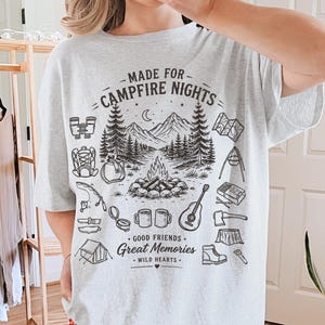 May include: A light gray t-shirt with a black graphic design featuring a campfire scene, mountains, and camping-related icons. The text "MADE FOR CAMPFIRE NIGHTS" and "GOOD FRIENDS, GREAT MEMORIES, WILD HEARTS" are also printed on the shirt.