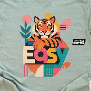 Pode incluir: Uma t-shirt de manga curta em verde s&aacute;lvia claro com uma ilustra&ccedil;&atilde;o gr&aacute;fica de um tigre com riscas cor de laranja, pretas e brancas. O design inclui a palavra "EASY" em letras grandes e coloridas, juntamente com formas geom&eacute;tricas e motivos vegetais.