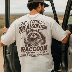 May include: An off-white short-sleeve t-shirt featuring a dark brown graphic on the back. The design shows a raccoon in a trash can with the text: STOP ASKING THE ALGORITHM ASK A RACCOON I ALSO MAKE STUFF UP AND I HAVE TINY HANDS.