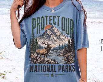 May include: A blue jean colored t-shirt featuring a graphic print of a mountain range, a forest, a river, and an elk. The text reads PROTECT OUR NATIONAL PARKS. The design includes a sun and pine trees in shades of brown, green, and cream.