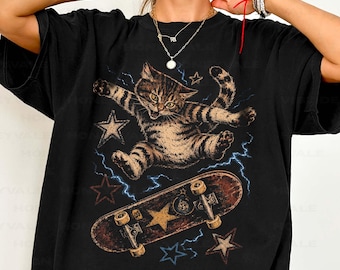 May include: A black short-sleeve t-shirt featuring a graphic print of a brown tabby cat jumping above a skateboard. The design includes blue lightning bolts and several stars in various sizes. The word BLACK is printed on the right side of the shirt.