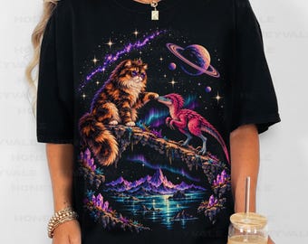 May include: A black t-shirt featuring a graphic print of a fluffy brown cat and a pink dinosaur touching noses on a rocky cliff. The background displays a cosmic scene with a planet, stars, purple mountains, and a glowing aurora borealis.