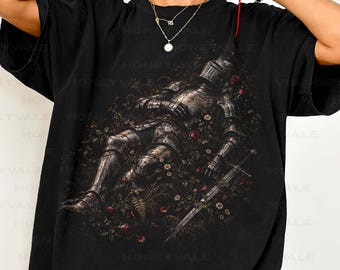 May include: A black short-sleeve t-shirt featuring a graphic print of a knight in full metal armor lying down amidst a field of dark red roses and small white flowers, with a long sword resting on the ground beside the armored figure.