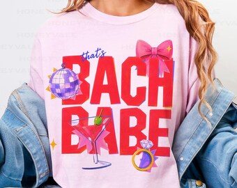 May include: A light pink t-shirt featuring the red text that's BACH BABE. The design includes a purple disco ball, a pink martini glass with a green olive, a pink bow, and a gold diamond ring. The shirt is worn under a blue denim jacket.