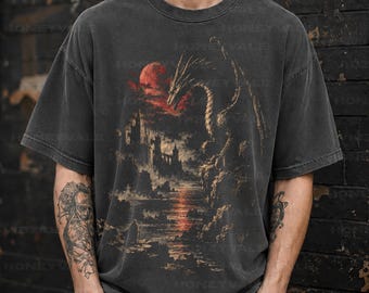 May include: A charcoal grey short-sleeve t-shirt featuring a graphic print of a dragon perched on a cliff overlooking a castle and a red moon. The text Comfort Colors PEPPER is printed in the upper right corner of the garment.