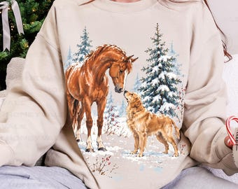 May include: A sand-colored crewneck sweatshirt featuring a graphic print of a brown horse and a golden dog standing in a snowy forest with pine trees. The text "Gildan 18000 SAND" is printed in the upper right corner of the image.