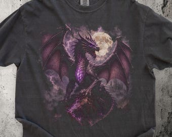 May include: A charcoal gray short-sleeve t-shirt featuring a graphic print of a purple dragon with large wings perched on a rock against a full moon and stormy sky. The shirt has a crew neckline and the text Comfort Colors PEPPER.