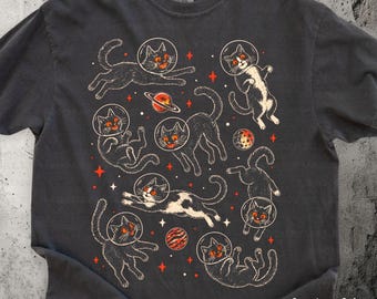 May include: A charcoal gray t-shirt featuring a graphic print of multiple cats wearing glass space helmets, floating among orange and white stars and planets. The shirt includes the text Comfort Colors PEPPER in the bottom right corner.