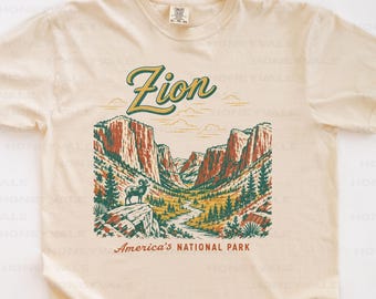 May include: A cream-colored short-sleeve t-shirt featuring a graphic of Zion National Park. The design includes a bighorn sheep on a rock, canyon walls, trees, and a winding path. The text reads Zion and America's NATIONAL PARK.