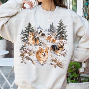 May include: A sand-colored crewneck sweatshirt featuring a graphic print of multiple Corgi dogs in a snowy forest setting with pine trees. The dogs are depicted in various poses amidst snow-covered ground and dry winter grasses.