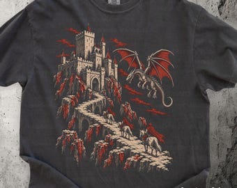 May include: A charcoal gray short-sleeve t-shirt featuring a graphic print of a stone castle on a mountain peak, a flying red-winged dragon, and figures on horseback traveling along a winding path. The shirt has a crew neckline.