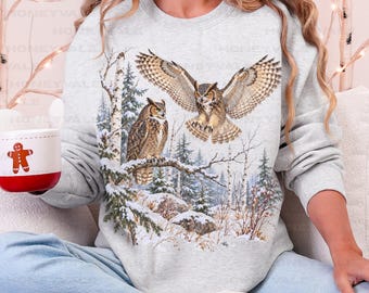 May include: A light gray crewneck sweatshirt featuring a graphic print of two owls in a snowy forest setting, with one owl perched on a branch and the other in flight. The sweatshirt is paired with light blue pants and white slippers.