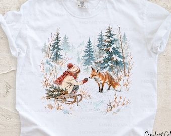 May include: A white short-sleeve t-shirt featuring a graphic illustration of a person in a winter coat and red scarf kneeling in the snow near a wooden sled, reaching toward a fox in a snowy forest setting with pine trees.