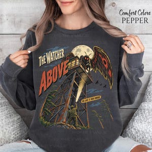 May include: A charcoal gray crewneck sweatshirt featuring a graphic print of a person standing at the base of a fire lookout tower, shining a flashlight upward at a large moth-like creature. The text reads The Watcher Above and No Light Is High Enough.