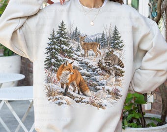 May include: A sand-colored crewneck sweatshirt featuring a detailed graphic print of a winter forest scene. The illustration depicts a red fox, a deer, a raccoon, and two rabbits in a snowy landscape with pine trees and a flowing stream.