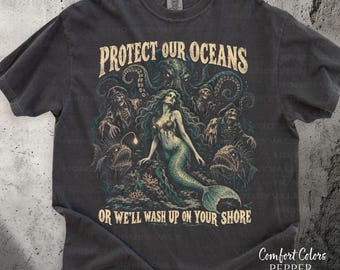 May include: A charcoal gray t-shirt featuring a graphic print of a mermaid, skeletons, and an octopus with the text "PROTECT OUR OCEANS OR WE'LL WASH UP ON YOUR SHORE" in a beige font. The shirt has a crew neckline and short sleeves.