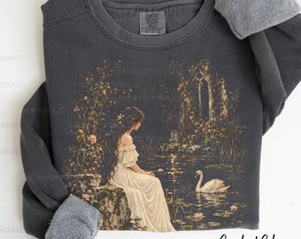 May include: A charcoal gray crewneck sweatshirt with a fleece interior. The front features a graphic of a person in a long cream-colored dress sitting on a stone ledge by a pond with a white swan, surrounded by climbing roses and stone arches.