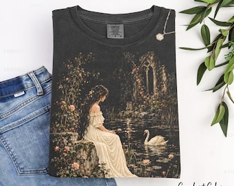 May include: A charcoal gray t-shirt featuring a graphic print of a person in a long cream dress sitting by a pond with a white swan, surrounded by stone ruins and climbing roses. The shirt has a Comfort Colors label and a small gold necklace.