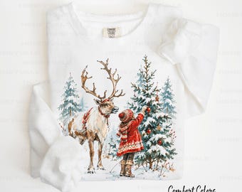 May include: A white long-sleeve sweatshirt featuring a watercolor graphic of a person in a red coat and hat decorating a snow-covered pine tree next to a reindeer. The inner neck label reads Comfort Colors.
