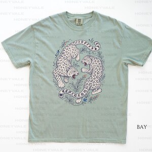May include: A sage green short-sleeve t-shirt featuring a central graphic illustration of two snow leopards in a circular arrangement surrounded by delicate botanical line art. The leopards are white with black spots and are depicted in a dynamic pose.