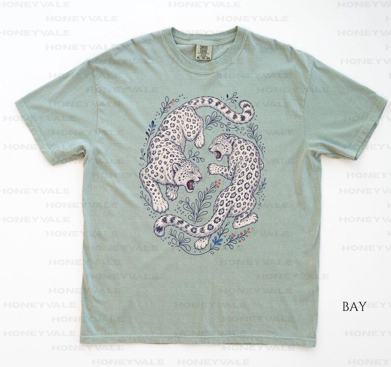 May include: A sage green short-sleeve t-shirt featuring a central graphic illustration of two snow leopards in a circular arrangement surrounded by delicate botanical line art. The leopards are white with black spots and are depicted in a dynamic pose.