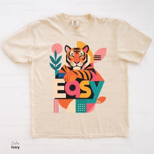 Pode incluir: Uma t-shirt de manga curta na cor marfim com uma estampa gr&aacute;fica de um tigre apoiando as patas na palavra "EASY" em letras grandes e coloridas. O design inclui formas geom&eacute;tricas, uma planta estilizada e um padr&atilde;o de pontos.
