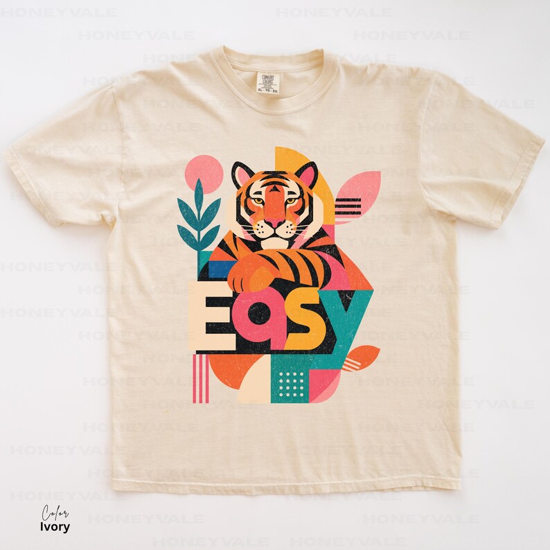 Pode incluir: Uma t-shirt de manga curta na cor marfim com uma estampa gr&aacute;fica de um tigre apoiando as patas na palavra "EASY" em letras grandes e coloridas. O design inclui formas geom&eacute;tricas, uma planta estilizada e um padr&atilde;o de pontos.