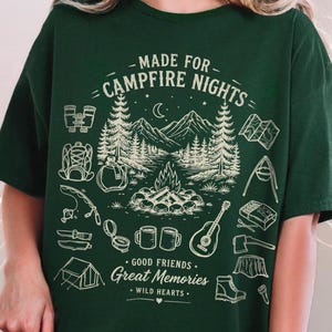 May include: A dark green t-shirt featuring a cream-colored graphic of a campfire scene with mountains, trees, and camping gear. The text reads: MADE FOR CAMPFIRE NIGHTS, GOOD FRIENDS, Great Memories, and WILD HEARTS, along with icons of a tent, guitar, and boots.