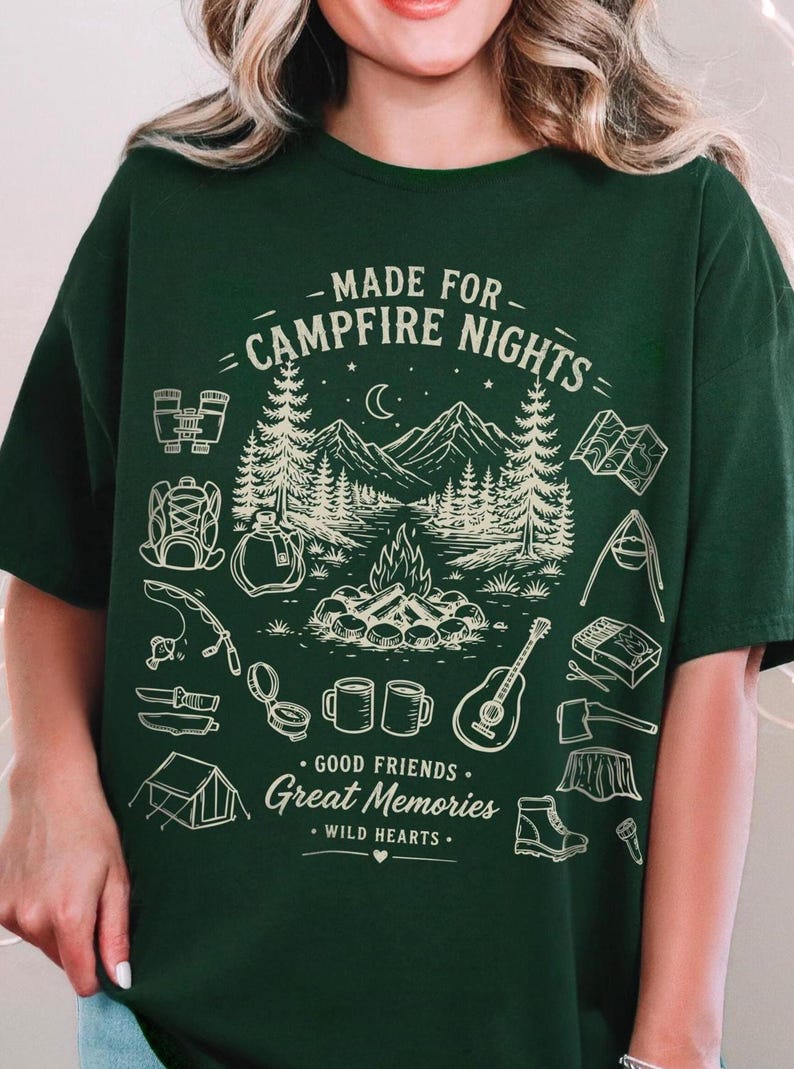 May include: A dark green t-shirt featuring a cream-colored graphic of a campfire scene with mountains, trees, and camping gear. The text reads: MADE FOR CAMPFIRE NIGHTS, GOOD FRIENDS, Great Memories, and WILD HEARTS, along with icons of a tent, guitar, and boots.