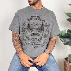 May include: A heather gray short-sleeve t-shirt featuring a black graphic print of a campfire scene with mountains, trees, and camping gear. The text reads MADE FOR CAMPFIRE NIGHTS, GOOD FRIENDS, GREAT MEMORIES, and WILD HEARTS in a stylized font.