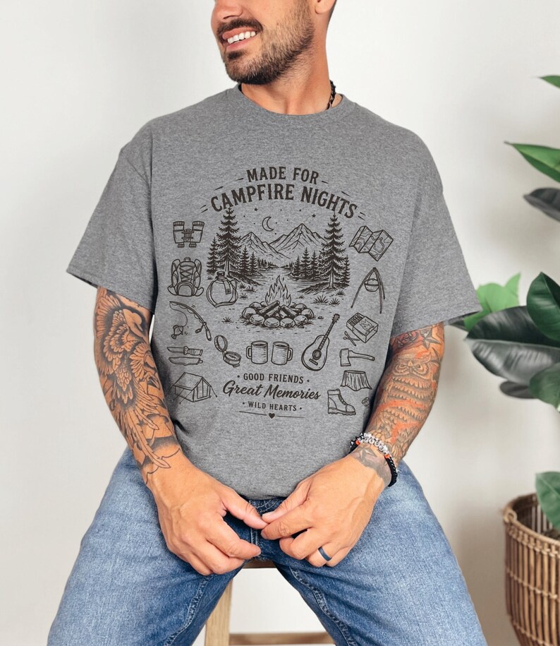 May include: A heather gray short-sleeve t-shirt featuring a black graphic print of a campfire scene with mountains, trees, and camping gear. The text reads MADE FOR CAMPFIRE NIGHTS, GOOD FRIENDS, GREAT MEMORIES, and WILD HEARTS in a stylized font.