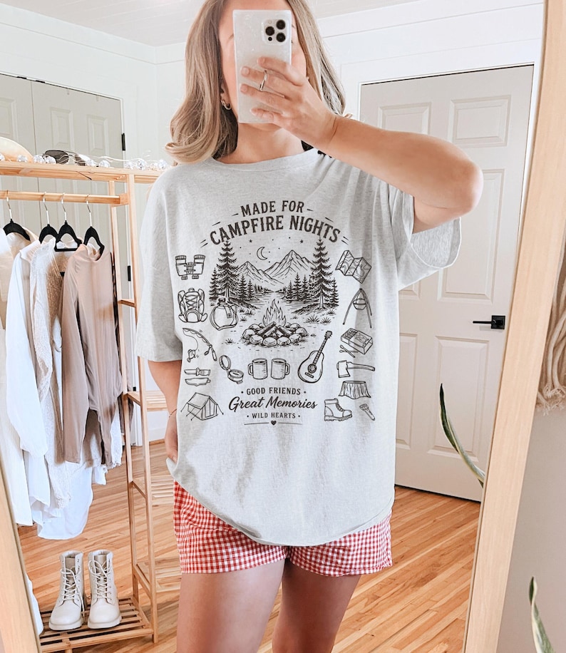 May include: A light gray t-shirt with a black graphic design featuring a campfire scene, mountains, and camping-related icons. The text "MADE FOR CAMPFIRE NIGHTS" and "GOOD FRIENDS, GREAT MEMORIES, WILD HEARTS" are also printed on the shirt.