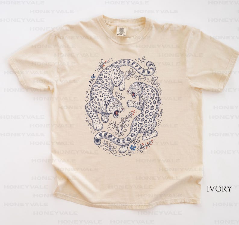May include: An ivory short-sleeve t-shirt featuring a central graphic illustration of two leopards in a circular arrangement surrounded by botanical elements. The leopards are depicted in dark blue ink with detailed spots and open mouths.