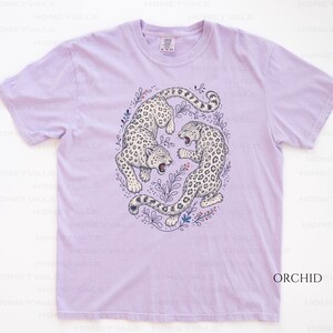 May include: A light purple short-sleeve t-shirt featuring a central graphic of two white leopards with black spots in a circular arrangement. The leopards are surrounded by delicate line-drawn botanical elements in blue and red ink.