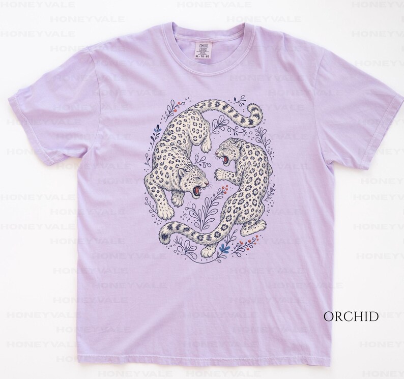 May include: A light purple short-sleeve t-shirt featuring a central graphic of two white leopards with black spots in a circular arrangement. The leopards are surrounded by delicate line-drawn botanical elements in blue and red ink.