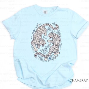 May include: A light blue short-sleeve t-shirt featuring a central graphic of two snow leopards in a circular arrangement surrounded by botanical illustrations. The leopards are depicted in white with black spots, and the text CHAMBRAY is visible.