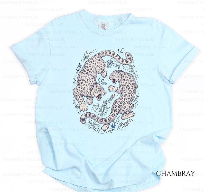 May include: A light blue short-sleeve t-shirt featuring a central graphic of two snow leopards in a circular arrangement surrounded by botanical illustrations. The leopards are depicted in white with black spots, and the text CHAMBRAY is visible.